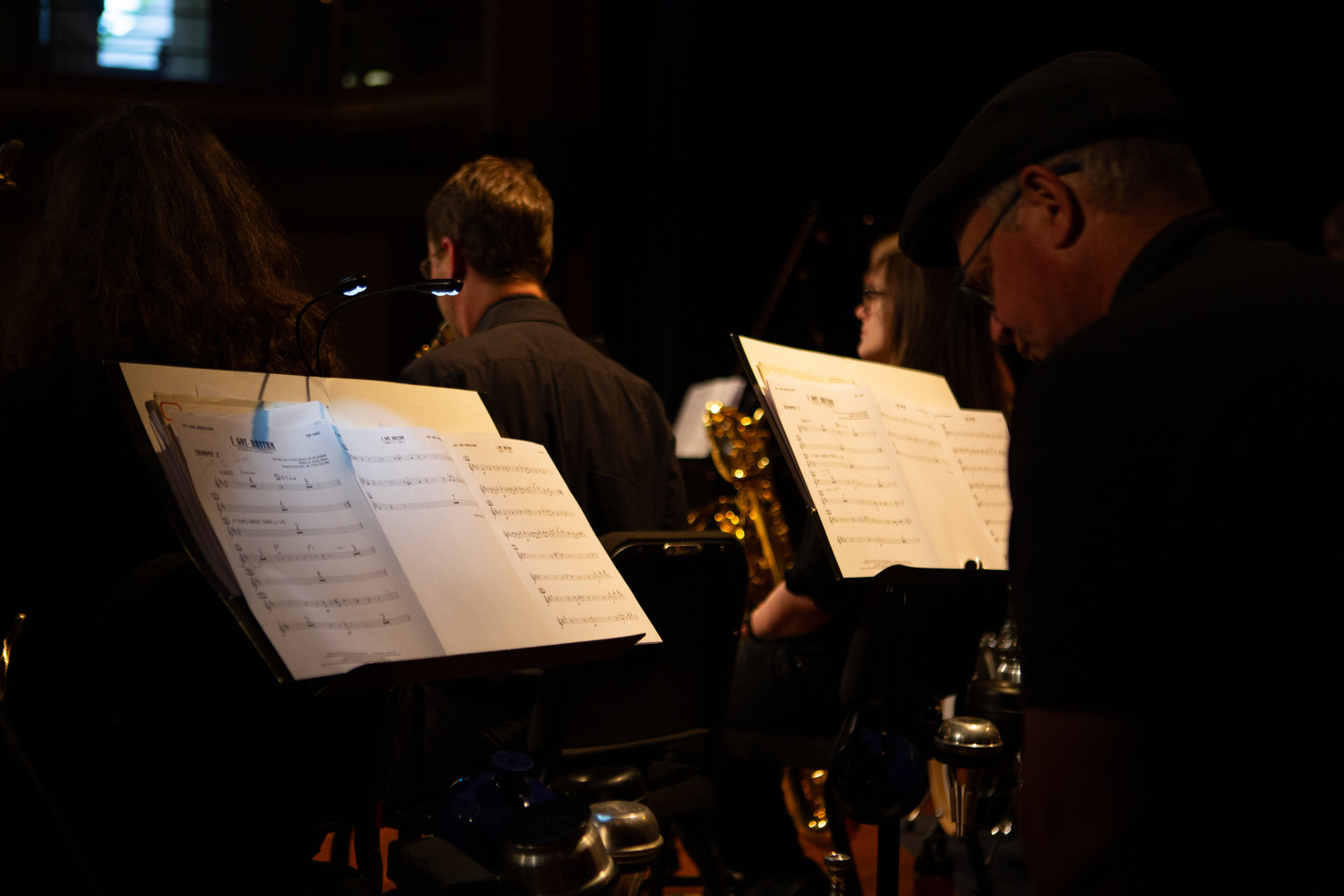 A view from behind the band of lit music stands and sheet music, brass glinting in the low stage light.