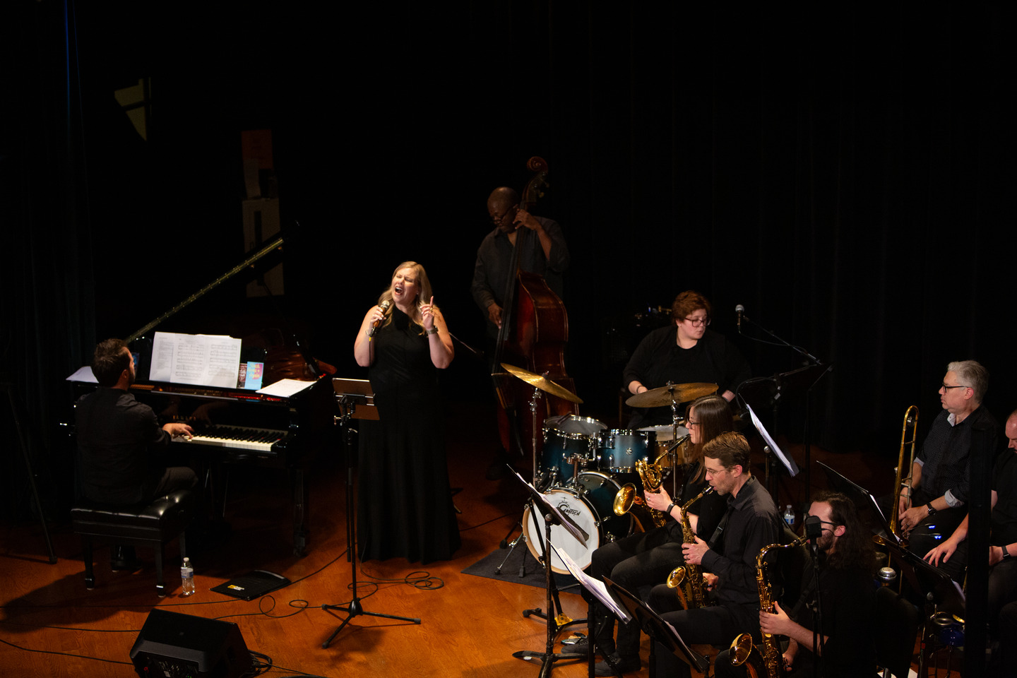The vocalist singing at center stage with the pianist, upright bassist, drummer, and horn section across the stage.