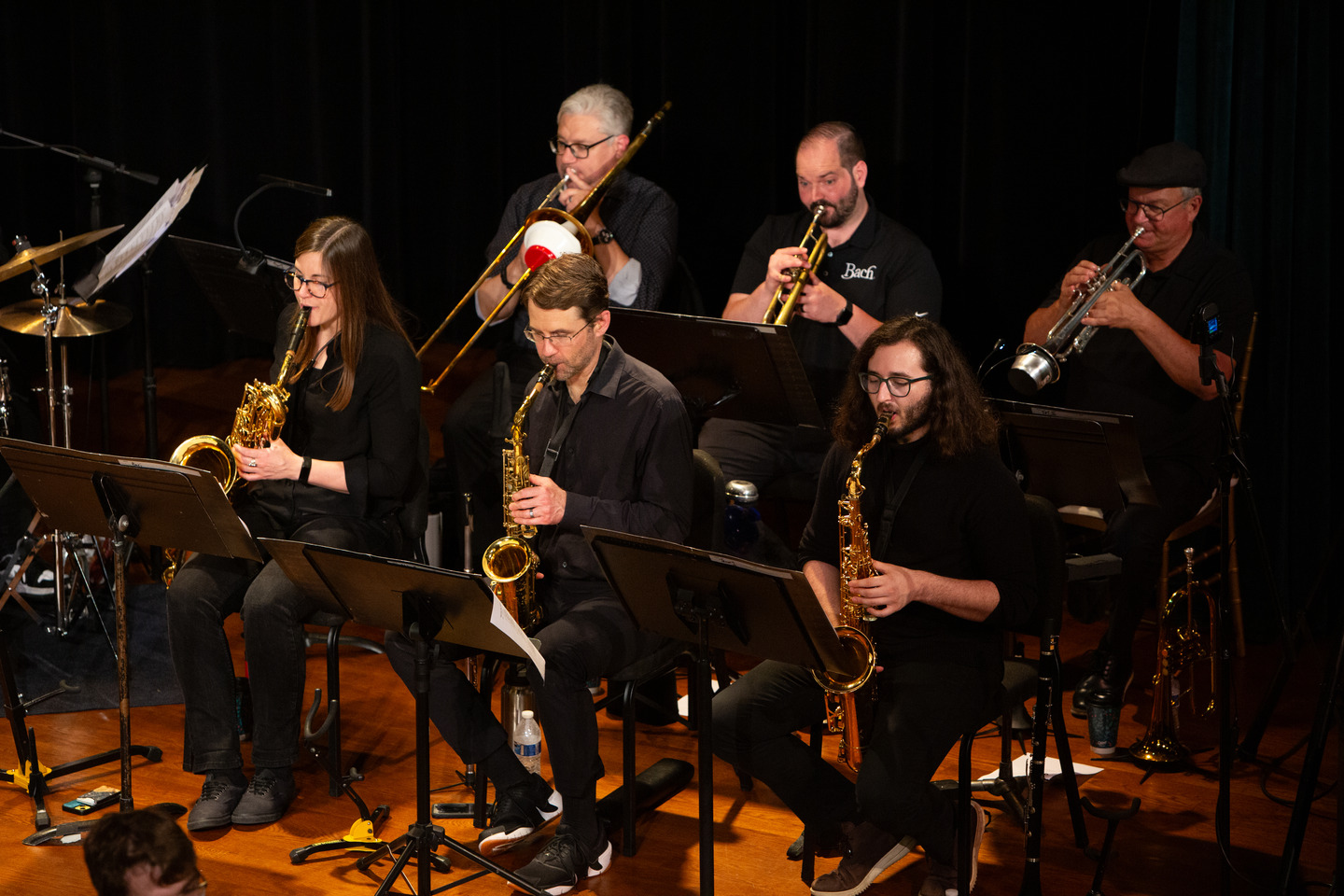 The full horn section seated in a row — three saxophones in front, trombone and trumpets behind.